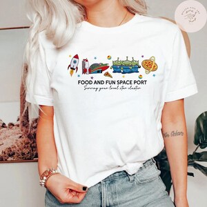 May include: White t-shirt featuring a colorful graphic of a rocket, spaceship, aliens, pizza, and stars. The text "FOOD AND FUN SPACE PORT" and "Serving your local star cluster" are printed below the graphic. The shirt is a casual, everyday wear item.