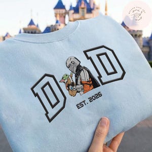 May include: Light blue sweatshirt with the word "DAD" in large, black, block letters. The design features an illustration of a Mandalorian character holding Grogu, with "EST. 2026" below. The sweatshirt is held by a hand.