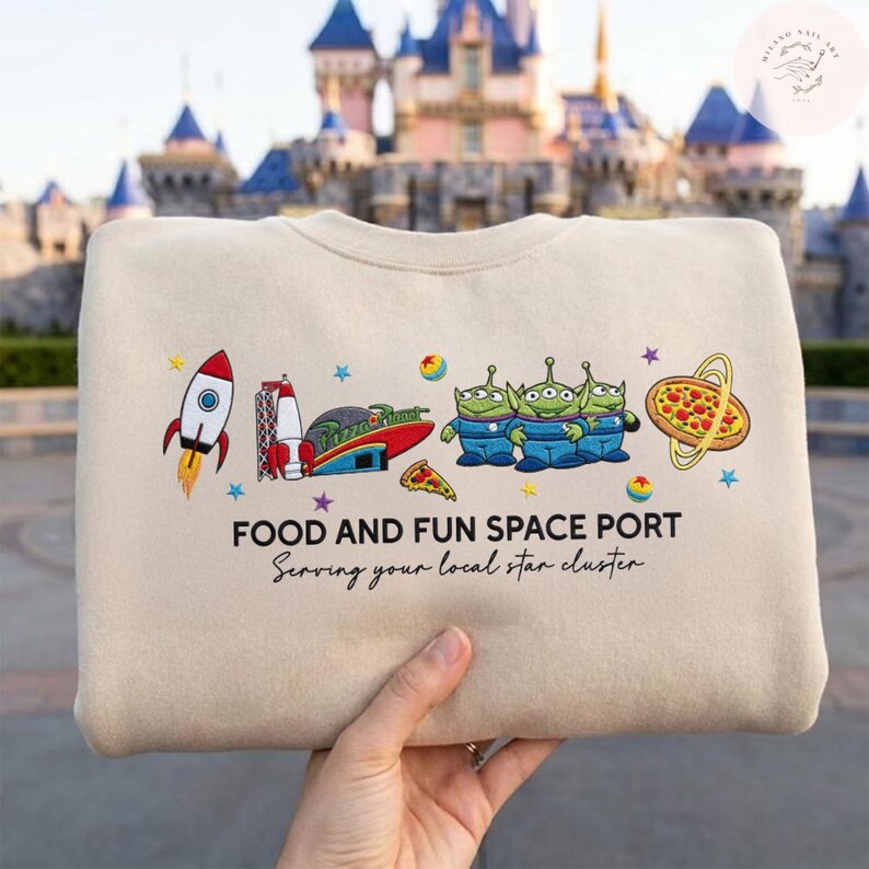 May include: Beige sweatshirt with a colorful graphic featuring a rocket, space port, pizza, and three green aliens. The text reads "FOOD AND FUN SPACE PORT" and "Serving your local star cluster."