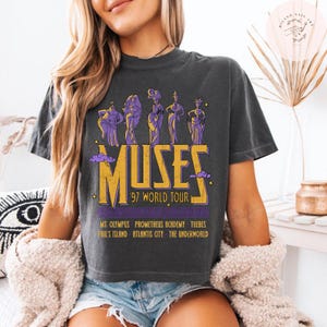 May include: Dark gray t-shirt featuring a graphic of the Muses with the text "MUSES '97 WORLD TOUR". The design includes purple figures and gold lettering. Additional text includes locations like Mt. Olympus and Atlantis City.