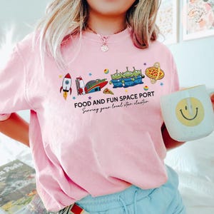 May include: Pink t-shirt with a colorful graphic featuring a rocket, spaceship, pizza, and three green aliens. The text reads "FOOD AND FUN SPACE PORT Serving your local star cluster."