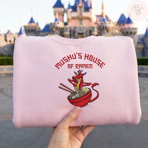 Op de afbeelding: Een lichtroze sweatshirt met de tekst "Mushu's House of Ramen" in het rood. Het ontwerp bevat een illustratie van Mushu de draak met eetstokjes boven een kom ramen. Het sweatshirt wordt voor een kasteel gehouden.