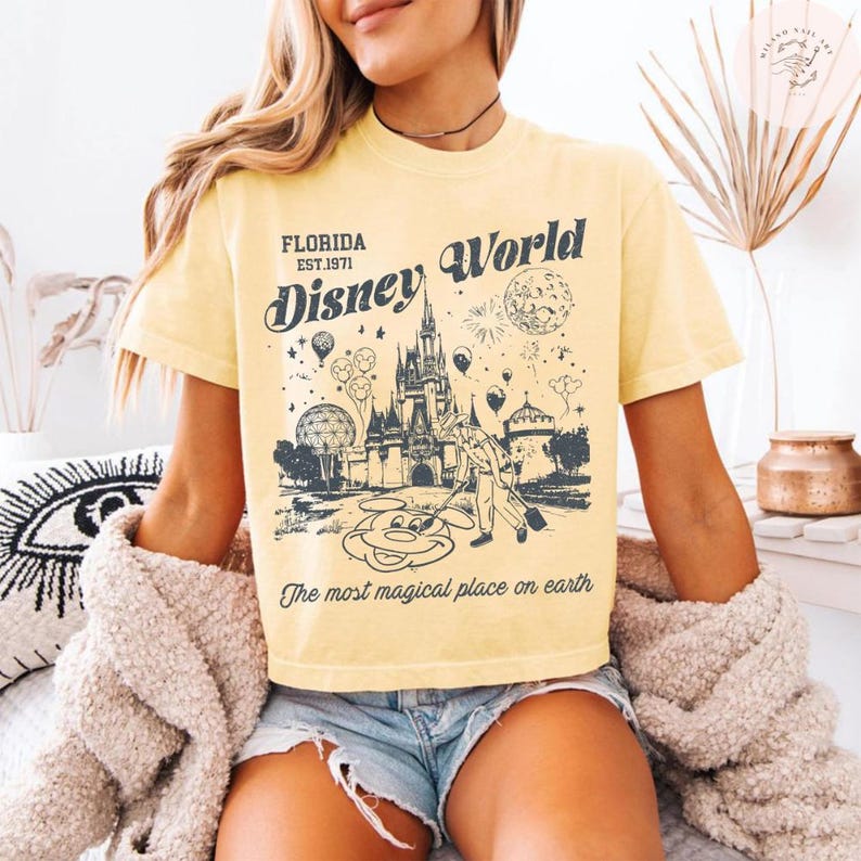 Może przedstawiać: Ż&oacute;łta koszulka z nadrukiem w stylu vintage z motywem Disney World. Projekt zawiera napisy "Florida Est. 1971" i "Disney World" z ilustracją zamku, Myszki Miki i balon&oacute;w. Tekst "The most magical place on earth" jest r&oacute;wnież zawarty.