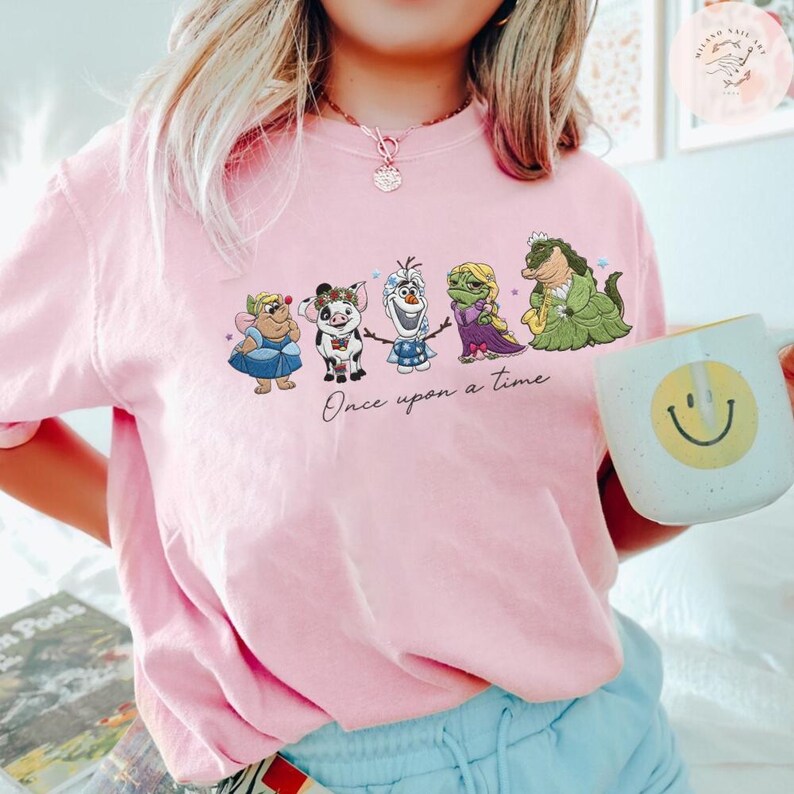 May include: Pink crew neck t-shirt featuring a cartoon graphic of animated characters with the text "Once upon a time". The characters include a fairy, pig, snowman, princess, and alligator. A white mug with a smiley face is held in the hand.
