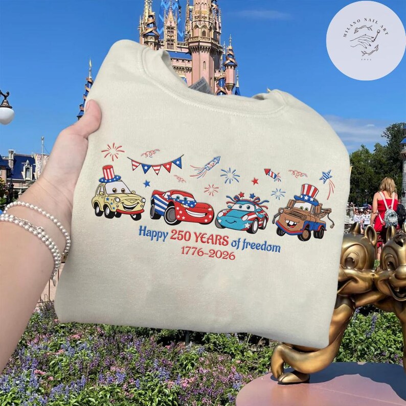 Pode incluir: Um moletom creme com carros de desenho animado com designs patri&oacute;ticos. Os carros s&atilde;o decorados com as cores vermelho, branco e azul e est&atilde;o dispostos em uma fileira. O texto "Happy 250 YEARS of freedom 1776-2026" est&aacute; impresso abaixo.