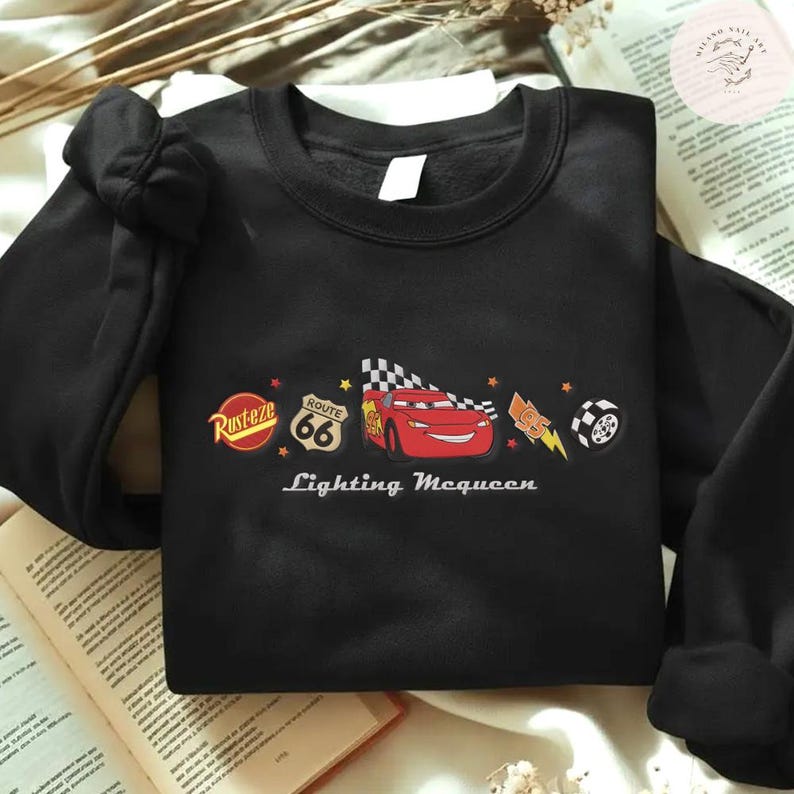May include: Black crew neck sweatshirt with a Lightning McQueen graphic from the Cars film. The design includes the Route 66 logo, a checkered flag, and the word "Rusteze".