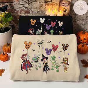 May include: A beige sweatshirt featuring an embroidered graphic of Toy Story characters in Halloween costumes holding Mickey-shaped balloons. Woody, Buzz Lightyear, Jessie, Rex, and Slinky Dog are depicted with festive details like capes and bandages.