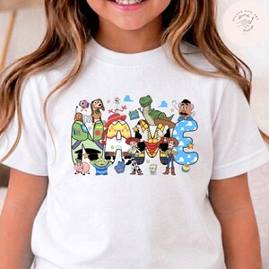 以下が含まれることがあります： 白い半袖Tシャツで、NAMEという文字の大きなグラフィックプリントが施されています。各文字は、バズ・ライトイヤー、ウッディ、ジェシー、レックス、ミスター・ポテトヘッドなど、トイ・ストーリーのキャラクターにインスパイアされた模様と色で塗りつぶされています。