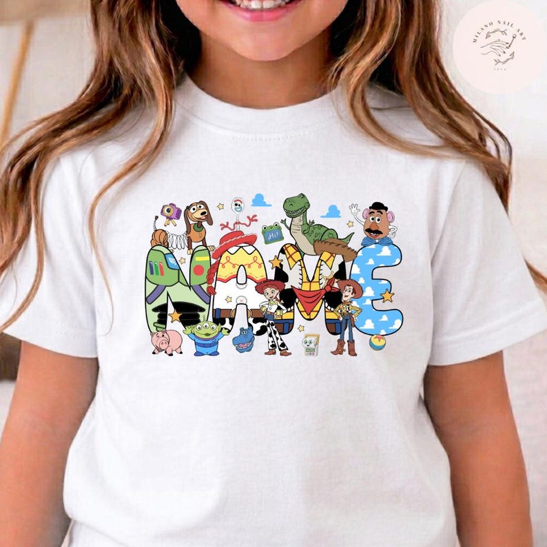 以下が含まれることがあります： 白い半袖Tシャツで、NAMEという文字の大きなグラフィックプリントが施されています。各文字は、バズ・ライトイヤー、ウッディ、ジェシー、レックス、ミスター・ポテトヘッドなど、トイ・ストーリーのキャラクターにインスパイアされた模様と色で塗りつぶされています。