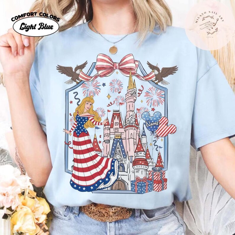 May include: Pale blue t-shirt with a patriotic design. It features a princess in a red, white, and blue dress, a castle, fireworks, and a Mickey Mouse balloon. The shirt has the words "Comfort Colors" and "Light Blue" printed on it.