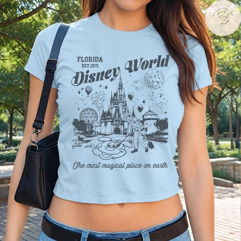 Może przedstawiać: Jasnoniebieski top z grafiką Disney World. Projekt zawiera zamek, Myszka Miki i tekst "Florida Est. 1971". Na koszulce wydrukowano r&oacute;wnież frazę "The most magical place on earth".