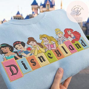 May include: Pale blue sweatshirt with an embroidered design of Disney princesses and the word "Disneyland" in colourful block letters. The design includes Jasmine, Snow White, Belle, Aurora, and Ariel. A castle is visible in the background.