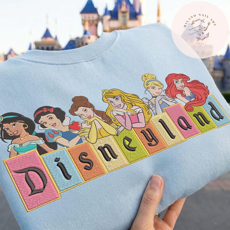May include: Pale blue sweatshirt with an embroidered design of Disney princesses and the word "Disneyland" in colourful block letters. The design includes Jasmine, Snow White, Belle, Aurora, and Ariel. A castle is visible in the background.