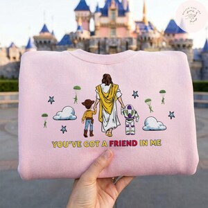 May include: Pale pink sweatshirt with a graphic of a figure in a white robe and yellow shawl holding hands with a cowboy and a space ranger. The text "YOU'VE GOT A FRIEND IN ME" is printed below the graphic.