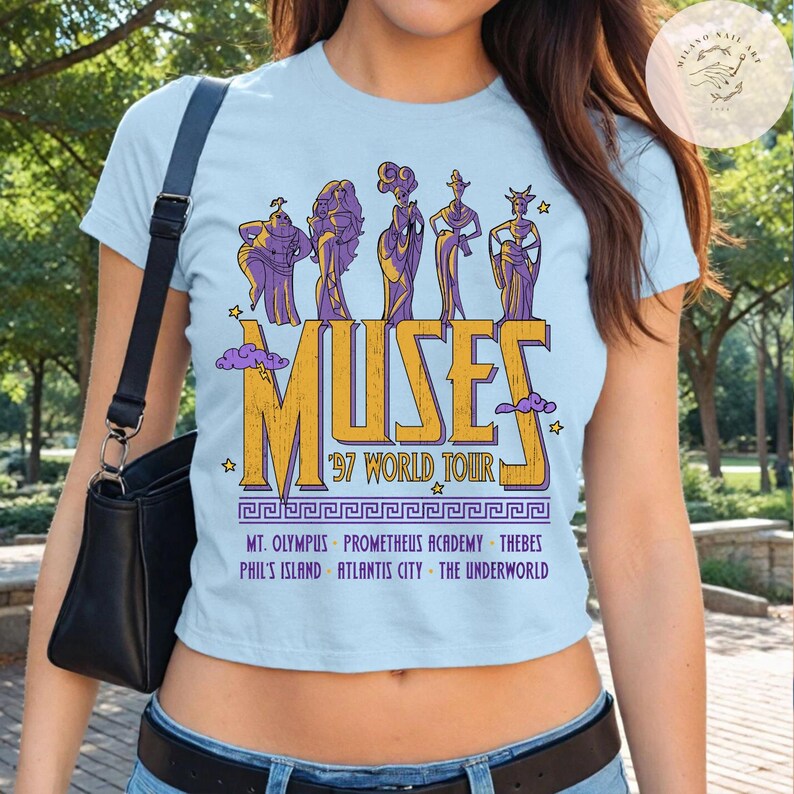 May include: Light blue crop top featuring a graphic of the Muses from the movie Hercules. The design includes the word "MUSES" in gold and purple, along with "'97 WORLD TOUR" and various locations. The shirt is a crew neck.