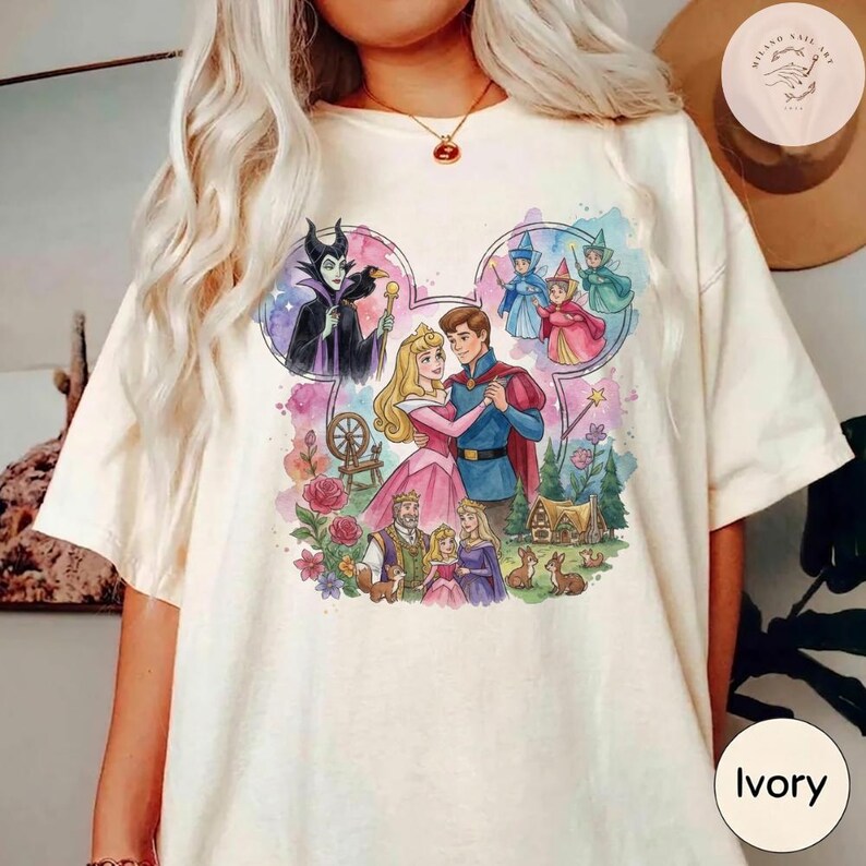May include: An ivory t-shirt featuring a colorful graphic print of characters from Sleeping Beauty arranged in a Mickey Mouse ear silhouette. The design includes Aurora, Prince Phillip, Maleficent, the three fairies, and forest animals.