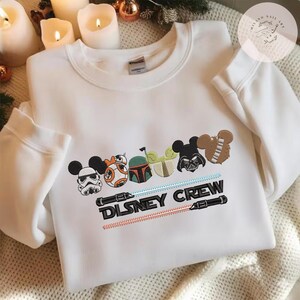 May include: White sweatshirt with embroidered Disney characters, such as Mickey Mouse, BB-8, and Darth Vader. The design includes lightsabers and the text "DISNEY CREW" in black.