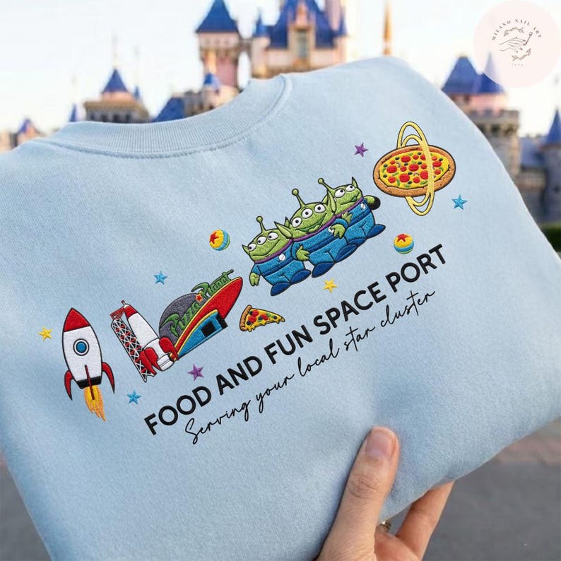 May include: Light blue sweatshirt with a colorful graphic featuring cartoon aliens, a rocket ship, and pizza. The text "FOOD AND FUN SPACE PORT" and "Serving your local star cluster" is printed below the graphic. The design is embroidered.