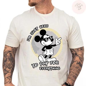 May include: Cream-colored t-shirt featuring a cartoon Mickey Mouse character wearing sunglasses with dollar signs. The text "I'M JUST HERE TO PAY FOR EVERYTHING" is printed above and below the character. The design is set against a gray and yellow circular background.