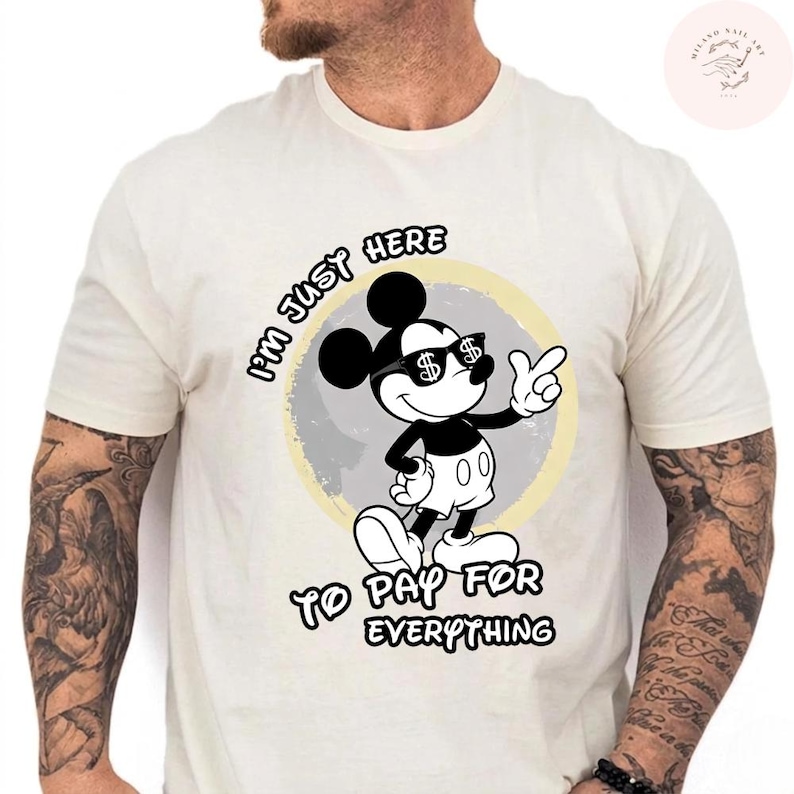 May include: Cream-colored t-shirt featuring a cartoon Mickey Mouse character wearing sunglasses with dollar signs. The text "I'M JUST HERE TO PAY FOR EVERYTHING" is printed above and below the character. The design is set against a gray and yellow circular background.