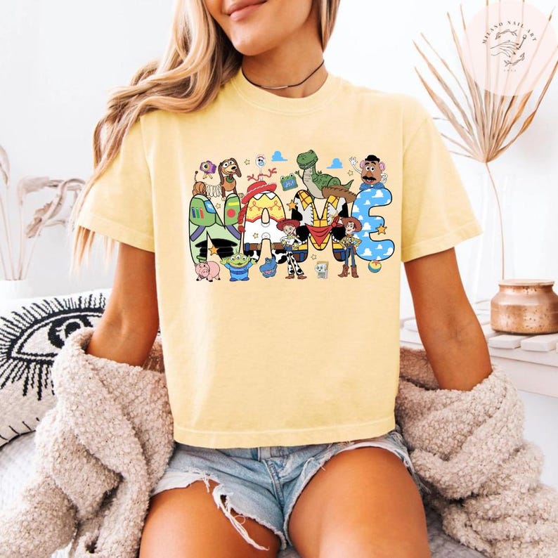 以下が含まれることがあります： 淡い黄色の半袖Tシャツで、NAMEという文字の大きなグラフィックプリントが施されています。各文字の中には、カウボーイ、スペースレンジャー、恐竜など、さまざまなアニメーションのおもちゃのキャラクターのカラフルなイラストが描かれています。