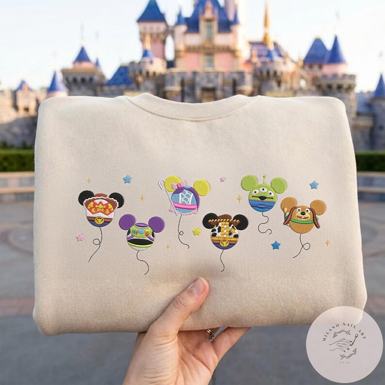 May include: Beige sweatshirt featuring embroidered Mickey Mouse-shaped balloons with various character designs. The balloons are in different colors and patterns, with blue stars embroidered around them. The background shows a castle.