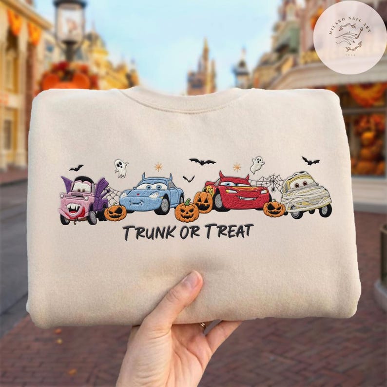 May include: A beige crewneck sweatshirt featuring an embroidered design of four cartoon cars dressed in Halloween costumes, including a vampire, a devil, and a mummy, alongside orange pumpkins, ghosts, bats, and the text TRUNK OR TREAT.