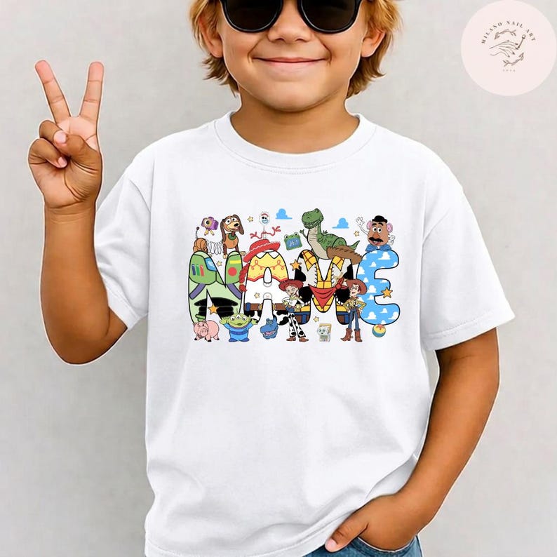 以下が含まれることがあります： 胸元にカラフルなグラフィックプリントが施された白い半袖Tシャツ。グラフィックには大きなブロック体で「NAME」という文字が描かれており、各文字はトイ・ストーリーの映画シリーズに登場する様々なキャラクターで装飾されています。