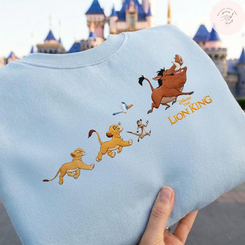 May include: Pale blue crew neck sweatshirt with an embroidered design from Disney's The Lion King. The design features Simba, Timon, Pumbaa, and Zazu. The words "The Lion King" are embroidered in gold. The sweatshirt is a casual style.
