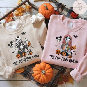 May include: A beige sweatshirt featuring a graphic of a skeleton character and the text THE PUMPKIN KING, and a light pink sweatshirt featuring a graphic of a female skeleton character and the text THE PUMPKIN QUEEN, both with pumpkins.