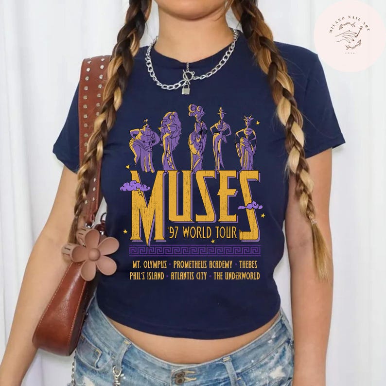 May include: Navy blue t-shirt featuring a graphic of the Muses from Greek mythology. The design includes the word "MUSES" in gold, the text "'97 WORLD TOUR", and various locations. The shirt has a classic crew neck.