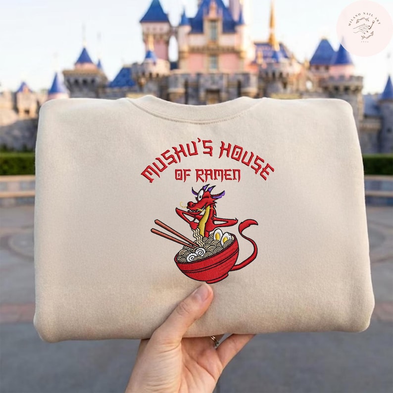 Op de afbeelding: Beige sweatshirt met de rode tekst "Mushu's House of Ramen". Daaronder een geborduurd beeld van Mushu de draak die stokjes boven een kom ramen vasthoudt. De achtergrond is wazig.