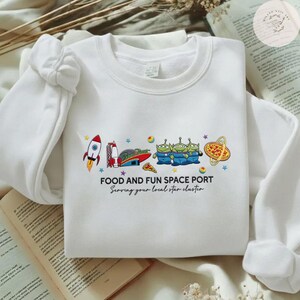 May include: White sweatshirt with a colorful graphic of a rocket, space aliens, pizza, and the text "FOOD AND FUN SPACE PORT". The text "Serving your local star cluster" is also included.