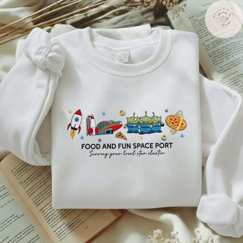 May include: White sweatshirt with a colorful graphic of a rocket, space aliens, pizza, and the text "FOOD AND FUN SPACE PORT". The text "Serving your local star cluster" is also included.