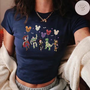 May include: A navy blue short-sleeved t-shirt featuring a graphic print of five cartoon characters dressed in Halloween costumes, each holding a Mickey Mouse-shaped balloon. The characters include a cowboy, a space ranger, a cowgirl, a dinosaur, and a dog.