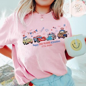 Pode incluir: Camiseta rosa claro com um gr&aacute;fico de carros de desenho animado decorados com temas patri&oacute;ticos, incluindo um carro com um chap&eacute;u do Tio Sam. O texto diz "Happy 250 Years of freedom 1776-2026". Uma caneca branca com um rosto sorridente &eacute; segurada.