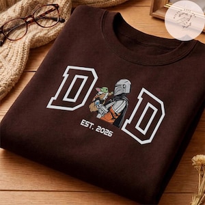 May include: A brown sweatshirt with the word "DAD" in white block letters. An illustration of a Mandalorian character holding Grogu is in the center. The text "EST. 2026" is below the illustration.