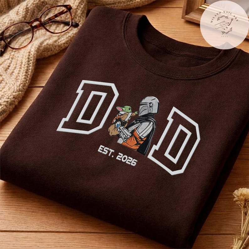 May include: A brown sweatshirt with the word "DAD" in white block letters. An illustration of a Mandalorian character holding Grogu is in the center. The text "EST. 2026" is below the illustration.