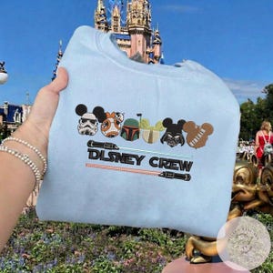 May include: Pale blue sweatshirt with embroidered Disney and Star Wars characters, including Mickey Mouse, Darth Vader, and Boba Fett. The text "DISNEY CREW" is embroidered below the characters, with lightsabers on either side.