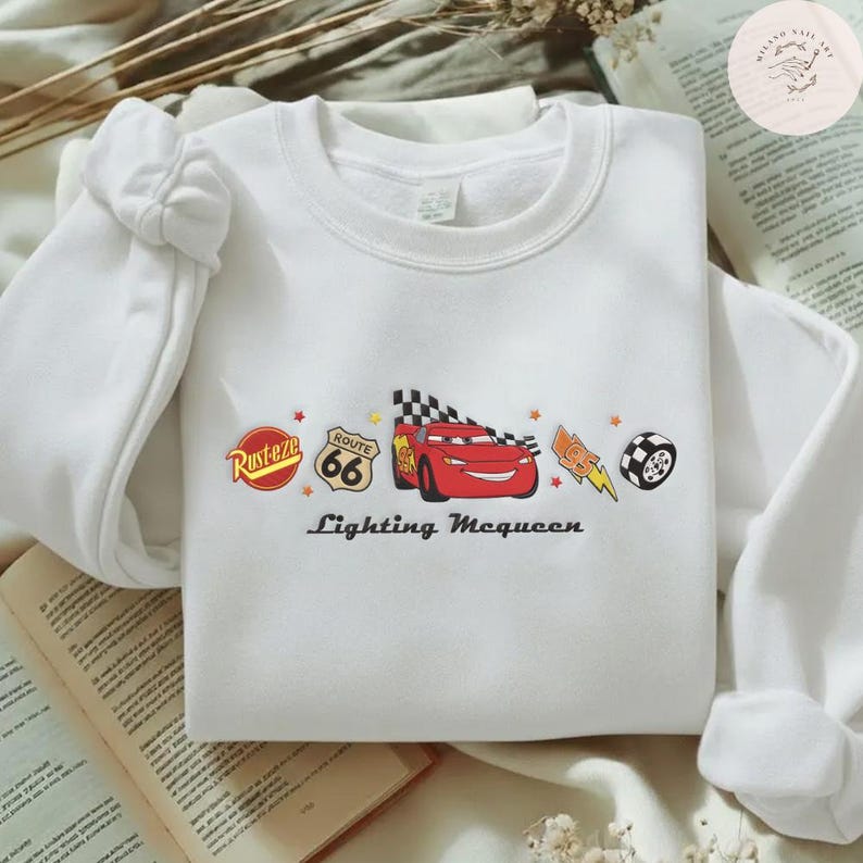 May include: White crew neck sweatshirt with a colourful graphic of Lightning McQueen from the Cars film. The design includes the Route 66 logo, a chequered flag, and the text "Lightning McQueen". The sweatshirt has long sleeves.
