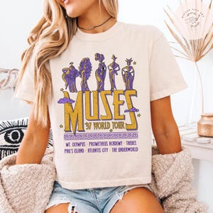 May include: Cream-colored t-shirt featuring a graphic of the Muses from the movie Hercules. The design includes the word "MUSES" in gold and purple, along with "'97 WORLD TOUR" and various locations.