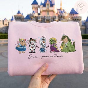 May include: A light pink sweatshirt featuring embroidered cartoon characters. The design includes a fairy, pig, snowman, frog, and alligator. The phrase "Once upon a time" is written below the characters. The background shows a castle.