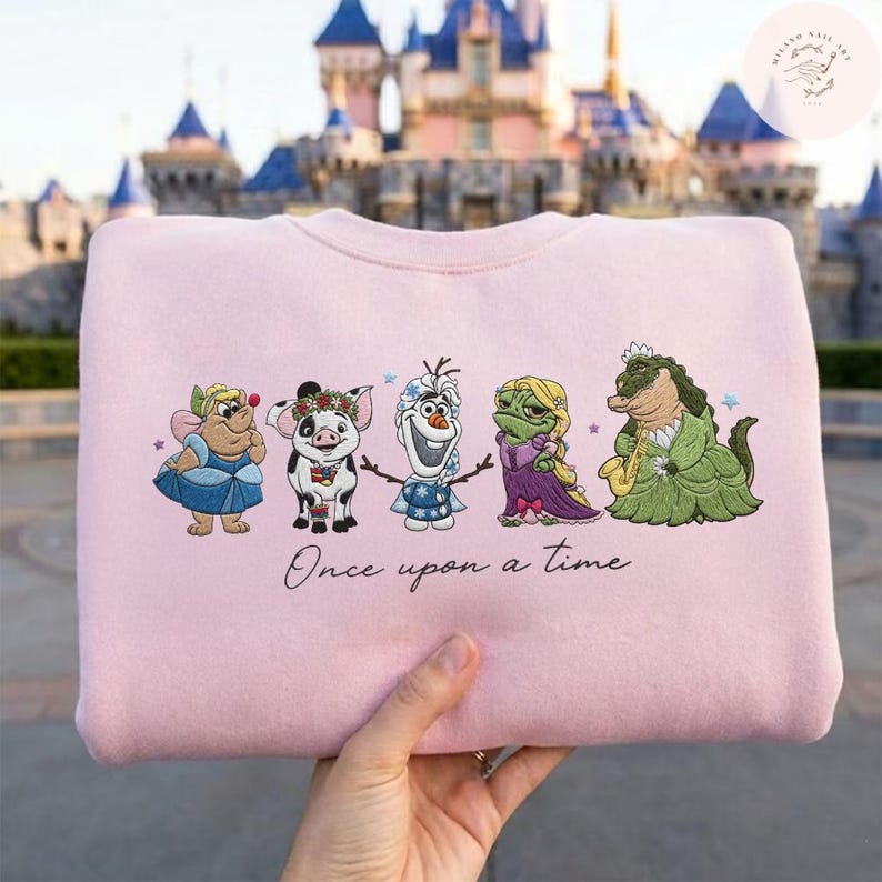 May include: A light pink sweatshirt featuring embroidered cartoon characters. The design includes a fairy, pig, snowman, frog, and alligator. The phrase "Once upon a time" is written below the characters. The background shows a castle.