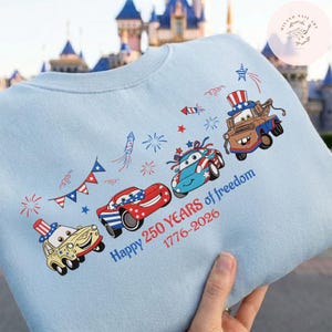 Pode incluir: Camisola azul clara com carros de desenho animado decorados com temas patri&oacute;ticos. O design inclui fogos de artif&iacute;cio, estrelas e o texto "Happy 250 Years of freedom 1776-2026". Os carros s&atilde;o vermelhos, azuis e amarelos.