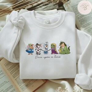May include: White sweatshirt featuring a colorful embroidered design of cartoon characters. The design includes a fairy, pig, snowman, princess, and alligator. The phrase "Once upon a time" is written below the characters.