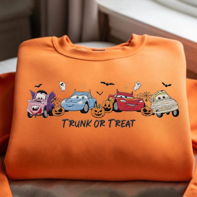 May include: An orange crewneck sweatshirt featuring an embroidered design of four cartoon cars dressed in Halloween costumes, including a vampire, devil, and mummy, surrounded by ghosts, bats, spiderwebs, and pumpkins with the text TRUNK OR TREAT.