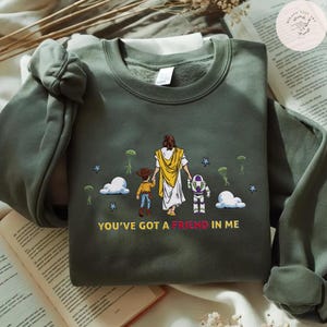 May include: Dark green sweatshirt with a graphic of a figure in a white robe and yellow shawl, holding hands with a cartoon cowboy and a space ranger. The text "YOU'VE GOT A FRIEND IN ME" is below the graphic.