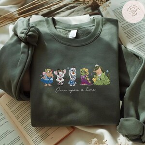 May include: Olive green sweatshirt featuring embroidered cartoon characters. The design includes a fairy, a pig, a snowman, a princess, and a crocodile. The phrase "Once upon a time" is written below the characters. The sweatshirt is folded on a book.