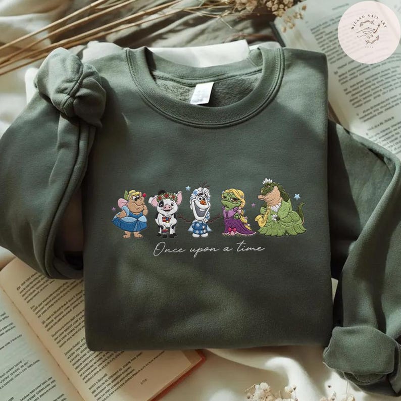 May include: Olive green sweatshirt featuring embroidered cartoon characters. The design includes a fairy, a pig, a snowman, a princess, and a crocodile. The phrase "Once upon a time" is written below the characters. The sweatshirt is folded on a book.