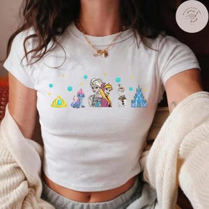 May include: White crop top featuring a colorful graphic of characters and elements from a popular animated movie. The design includes a crown, a blue dragon, two princesses, a snowman, and a castle.